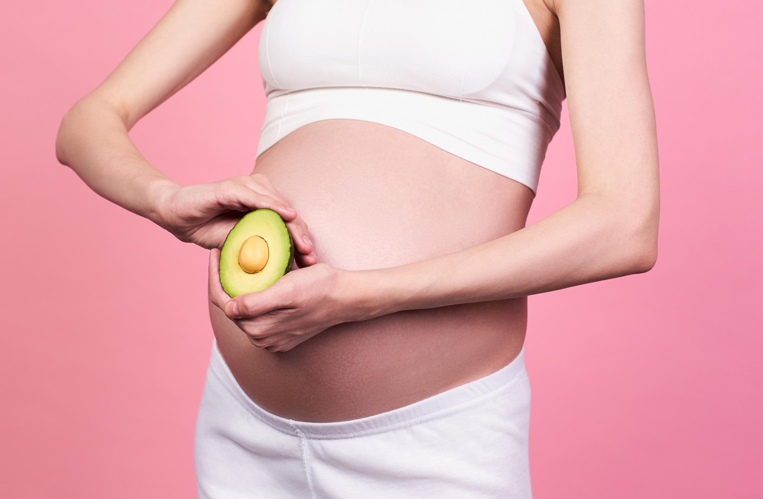 Close-up, profile of a young Caucasian woman, with beautiful healthy skin, in a white top, holding a half avocado at belly level with two hands, on a pink background. Pregnancy and nutrition concept.